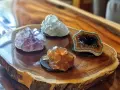 Cztery kryształy na drewnianej desce. Fioletowy ametyst, zielony kwarc, bursztynowy kalcyt i geoda z czarnymi kryształami. Opal, kamień znak zodiaku, może być jednym z nich.