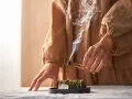 Dym z kadzidła unosi się, tworząc mistyczną atmosferę. Ręce trzymają drewno, symbolizując rytuały związane z magią i okultyzmem.