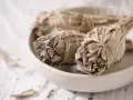 Trzy pęczki szałwii białej, związane sznurkiem, leżą w ceramicznej misie. Liście są suche, szaro-zielone, tworząc gęste, zwarte główki.