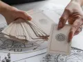 Dwie dłonie trzymają talię kart do wróżenia. Jedna ręka trzyma stos kart, druga wybiera jedną, by postawić tarota plus.