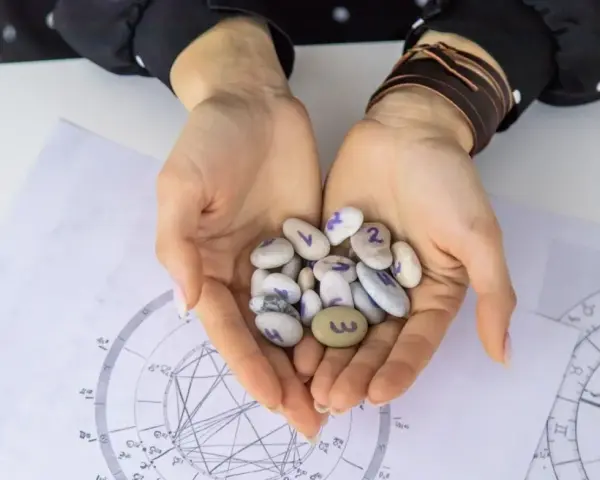 Dłonie trzymają kamyki z numerami, jeden z nich to 3, co może symbolizować numerologiczną 6. W tle wykresy astrologiczne.