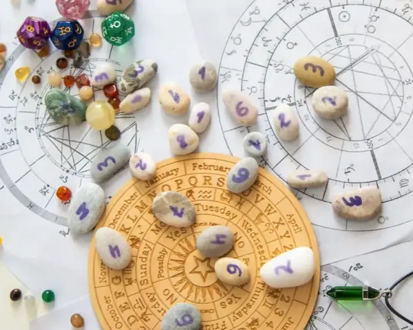 Kamienie z numerami, kostki do gry i talizmany na tle wykresów astrologicznych. Wibracja dnia 5 jest widoczna na jednym z kamieni.