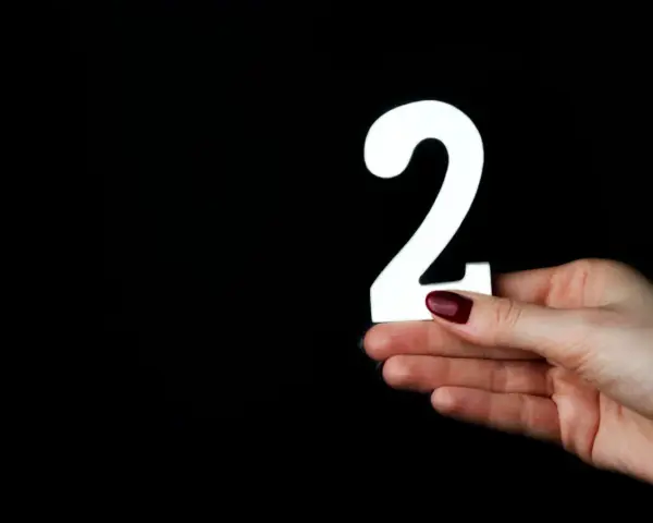 Dłoń z ciemnoczerwonym lakierem trzyma białą cyfrę 2. Co oznacza liczba 2?