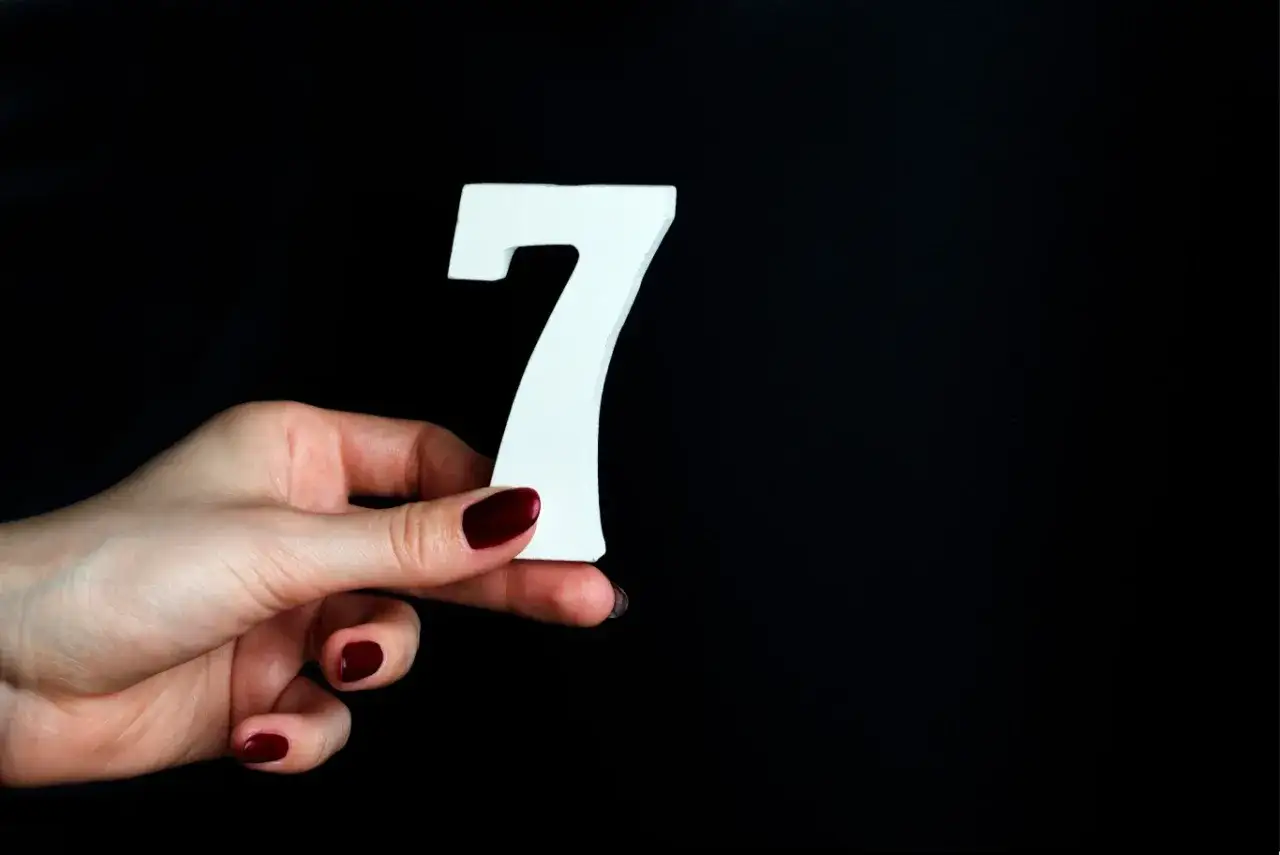 Dłoń z ciemnoczerwonym lakierem trzyma białą cyfrę 7, symbolizującą anielską liczbę 7.